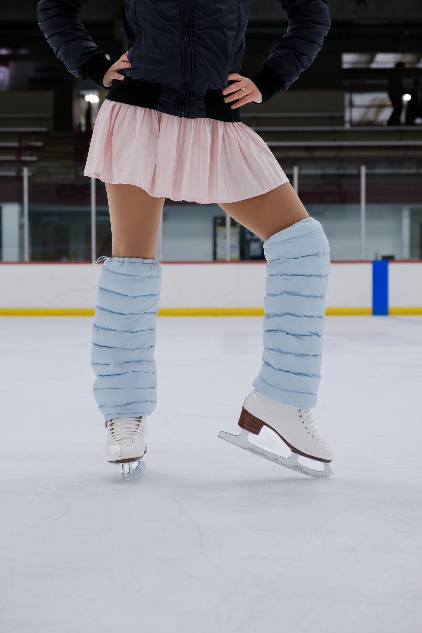 Person ice skating on an indoor rink wearing a pink skirt and light blue puffer leggings.