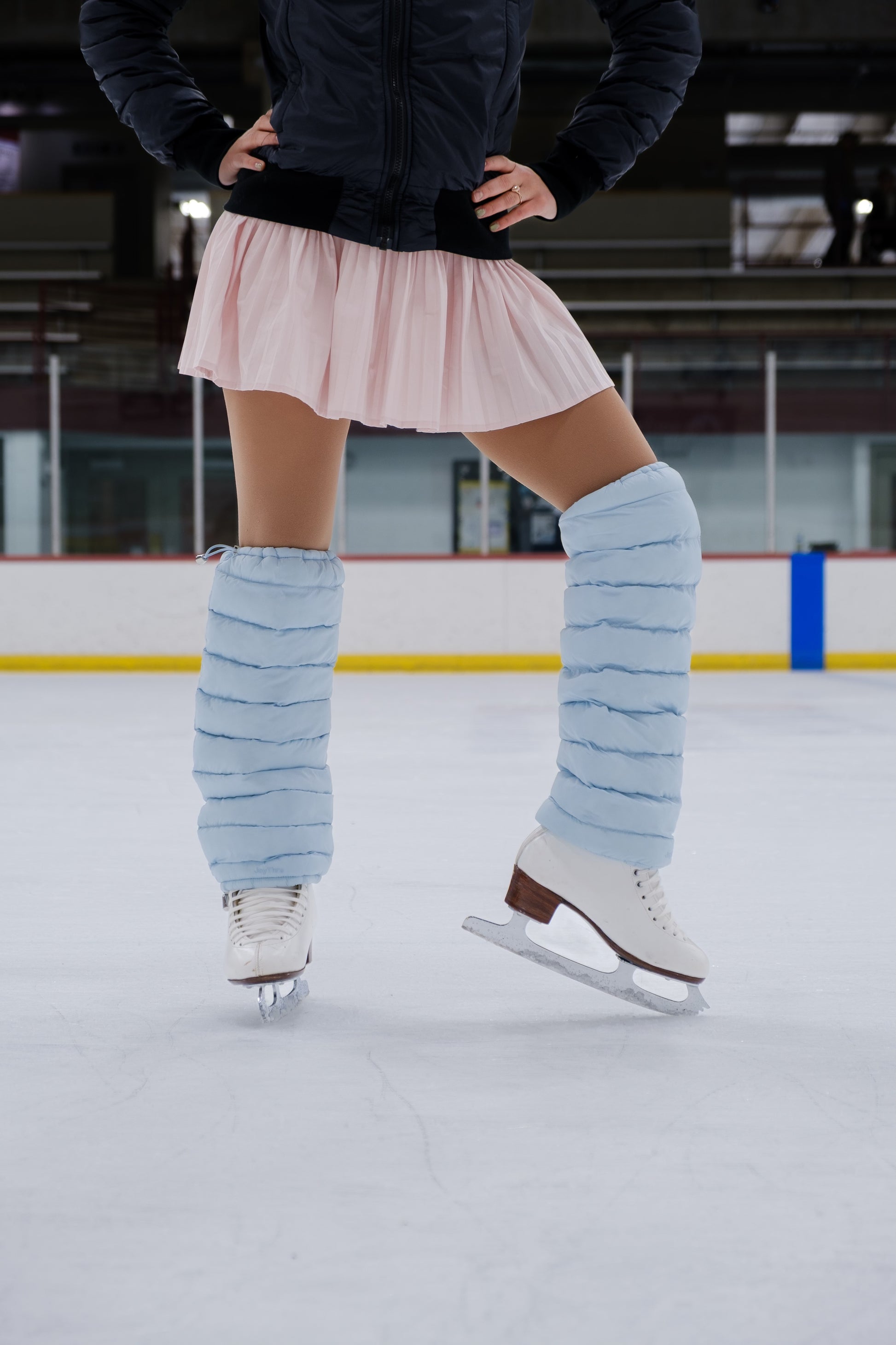 Person ice skating on an indoor rink wearing a pink skirt and light blue puffer leggings.